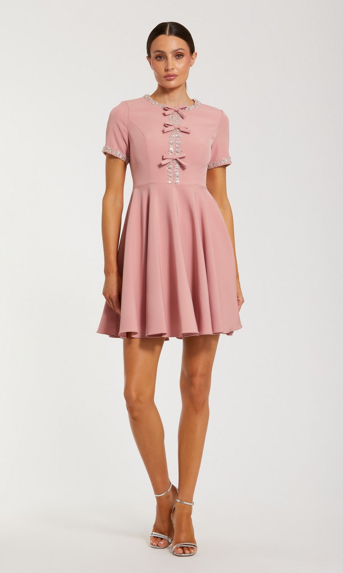 A woman stands facing forward in the Short Party Dress: Mac Duggal 56243, a dusty rose fit and flare mini dress with short sleeves, decorative bows, and lace trim, paired with silver high heels.
